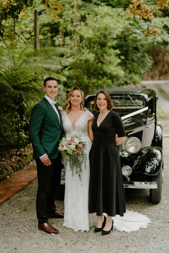 Eloise and Adam with wedding celebrant Charis White beside vintage car in lush garden setting
